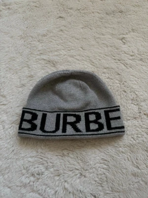 Grå Burberry mössa  - Säljer en grå stickad mössa från Burberry med bred svart logotyp runt hela kanten. 