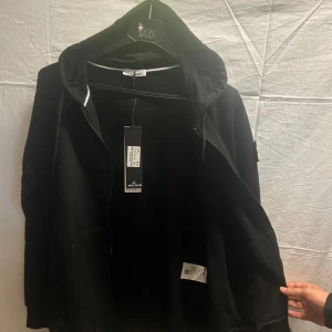 Svart hoodie med dragkedja från Stone Island - Svart hoodiejacka från Stone Island med dragkedja framtill, huva med snörning och klassisk logopatch på ärmen. Jackan har kängurufickor och är tillverkad i mjuk bomull. Perfekt för en clean och stilren streetlook.                Obs! Finns i alla storlekar. Skriv önskad storlek efter köp. 