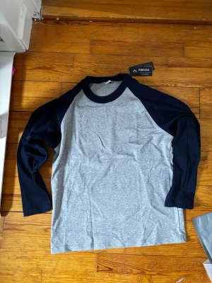 Grå och marinblå långärmad baseballtröja - Snygg långärmad baseballtröja i grått med marinblå ärmar och krage. Klassisk raglanmodell med kontrastfärgade detaljer. Tillverkad i mjukt bomullsmaterial som känns skönt mot huden. Perfekt för en avslappnad och sportig stil.