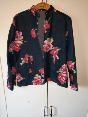 Blommig jeansjacka/denimjacka (Blå/Rosa) - Mörkblå blommig jeansjacka/denimjacka. Mönstrad med stora, livfulla rosor. Jackan har en knäppbar knapp vid kragen och knäppbara knappar vid ärmslutet. Storlek 42 på lappen, men passar dig som är en M/L. Perfekt som ett lättare ytterplagg till våren och sommaren eller som en snygg jacka inomhus över en enkel tröja.