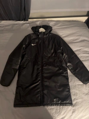 Svart lång parkas från Nike - Svart lång parkas med huva från Nike. Jackan har dragkedja framtill, två sidofickor och vit Nike-logga på bröstet. Baksidan har trycket 'stadium' i vitt. Materialet är glansigt och vattenavvisande, perfekt för kalla dagar.