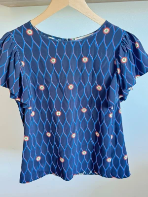 KENZO x H&M mörkblå blus, storlek 36 - S - En mönstrad blus från Kenzo x H&M kollektionen. Blusen har korta ärmar med volangdetaljer och en rund halsringning. Mönstret består av vågiga linjer i blått och rött samt små blommiga detaljer i vitt, gult och rött.  I fantastiskt fin skick kan inte så några skavanker  100% silke  Bröstvidd ca 43 cm Längd 58 cm