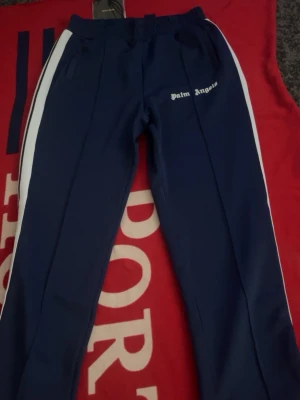 Blå Palm Angels mjukisbyxor - Säljer ett par blåa mjukisbyxor från Palm Angels med vita sidoränder och elastisk midja. Byxorna har raka ben och logga på vänster sida. Perfekta för en chill och sportig look. Pris kan diskuteras och mer bilder kan skickas