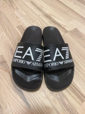 Svarta slides från Emporio Armani - Snygga svarta slides från Emporio Armani med stort vitt EA7-tryck och logga på remmen. Tillverkade i syntetmaterial med en klassisk form och bred rem över foten. Perfekta för en avslappnad och trendig stil.