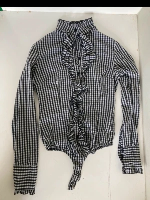 Italian coquette Vintage body blouse, s - Snygg svartvit rutig body med långa ärmar, hög krage och volangdetaljer längs knäppningen framtill. Kroppsnära passform och breda manschetter med volang. Perfekt för dig som gillar klassiska mönster med en twist.