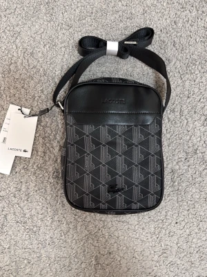 Svart axelväska från Lacoste - Snygg svart axelväska från Lacoste med justerbar rem och silverfärgade detaljer. Väskan har ett geometriskt mönster i grått och svart samt präglad Lacoste-logga på framsidan. Tillverkad i slitstarkt konstläder och har dragkedja upptill.