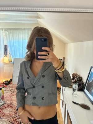 Grå cropped dubbelknäppt kavaj - Snygg grå cropped kavaj med dubbelknäppning och klassisk krage. Kavajen har fyra svarta knappar framtill och två dekorativa fickor. Passar perfekt till en trendig och avslappnad stil.