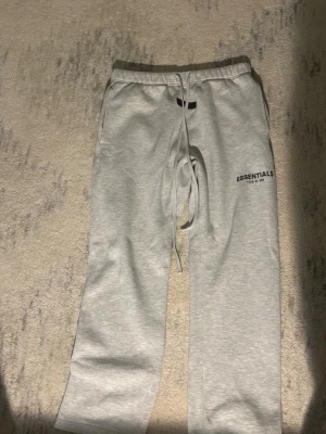 Gråa Essentials mjukisbyxor - Snygga ljusgrå mjukisbyxor från Fear of God Essentials med tryckt logga på benet. Byxorna har resår i midjan med dragsko och raka ben. Perfekta för en chill och avslappnad stil. Tillverkade i mjukt och bekvämt material.
