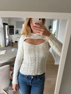 Vit kabelstickad cut out-tröja - Kabelstickad tröja med cut out-detalj över bröstet. En varm tröja som har lång ärm och en kroppsnära passform. Perfekt för dig som vill ha en unik twist på klassisk stickad stil.