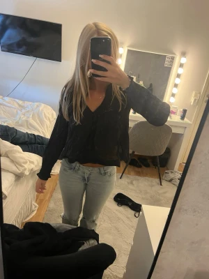 Svart v-ringad blus från Gina tricot  - Snygg svart blus med v-ringning och volangdetaljer vid axlarna. Blusen har långa ärmar och ett lätt, luftigt tyg som ger en avslappnad men ändå chic look. Perfekt att styla med jeans eller kjol för en trendig outfit.