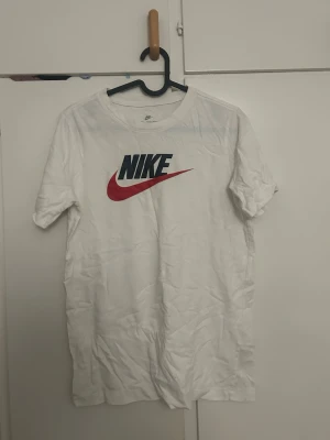 Vit Nike t-shirt med röd swoosh - Säljer en klassisk vit t-shirt från Nike med stor logga i svart och röd swoosh på bröstet. T-shirten är i mjuk bomull och har en normal passform med korta ärmar. Perfekt till jeans eller shorts för en sportig look.