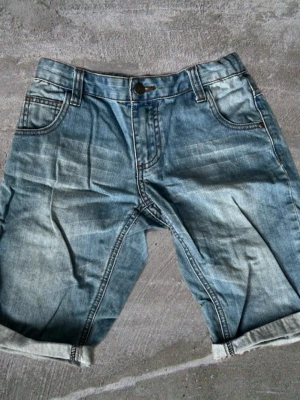 Blå jeansshorts  - Säljer ett par klassiska blå jeansshorts med upprullad kant och femficksmodell. Shortsen har normal passform, bälteshällor och knappgylf. Perfekta för sommaren och lätta att matcha med olika stilar.