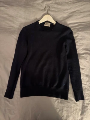 Mörkblå rundhalsad tröja - Snygg mörkblå tröja från Rowing Blazers med rund halsringning och diskret logga broderad nertill. Tröjan har långa ärmar och ribbade muddar vid ärmslut och nederkant. Perfekt för en stilren och enkel look.