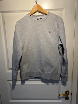 Grå sweatshirt från Lacoste - Klassisk grå sweatshirt från Lacoste med rund halsringning och den ikoniska gröna krokodilloggan broderad på bröstet. Tröjan har ribbade muddar vid ärmslut och nederkant. Perfekt för en clean och avslappnad stil.