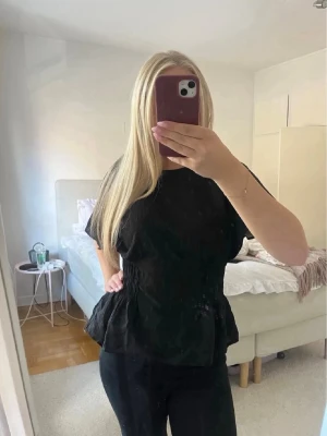 Svart blus med volangdetalj💓 - Säljer en svart blus med korta, lösa ärmar och volangdetalj nedtill. Blusen har en lätt rynkad midja som ger en snygg siluett och är tillverkad i ett tunt, luftigt material. Perfekt att styla med både jeans och kjol💓