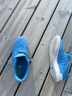 Blå Nike futsalskor med snörning - Säljer ett par ljusblå futsaldkor från Nike med vit sula och snörning. Skorna har quiltad ovandel och mönstrad yttersula för bra grepp på planen. Perfekta för dig som vill sticka ut på fotbollsplanen med stilrena och sportiga skor utan box. Pris kan diskuteras 
