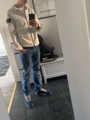 Beige overshirt från Stone Island - (PRISET EJ HUGGET I STEN) Snygg beige overshirt från Stone Island med dragkedja framtill och klassisk patch på vänster ärm. Overshirten har en bröstficka med lock och är tillverkad i ett lätt bomullsmaterial. Perfekt för lager-på-lager och streetwear vibes.