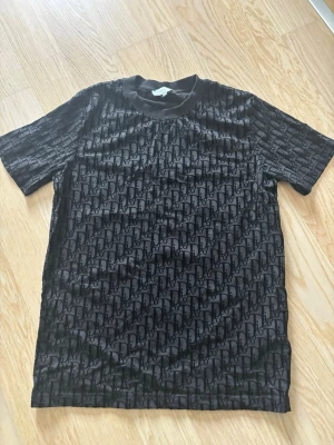 Svart t-shirt med mönster - Snygg svart t-shirt med all-over mönster i form av diskreta bokstäver. T-shirten har rund halsringning och korta ärmar. Materialet känns mjukt och följsamt, perfekt för en trendig och avslappnad look.