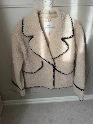 Krämvit teddyjacka från Object - Säljer en krämvit teddyjacka från Object med svart kontrastsöm längs kanterna. Jackan har en oversized krage, markerade sömmar och är gjord i mjukt, fluffigt material. Perfekt för dig som vill ha en mysig och trendig look. Endast testad!