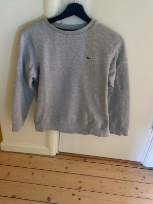 Lacost tröja 14år 164cm - Klassisk grå sweatshirt från Lacoste med rund halsringning och den ikoniska krokodilloggan broderad på bröstet. Tröjan har ribbade muddar vid ärmslut och nederkant. Perfekt för en avslappnad och stilren look.