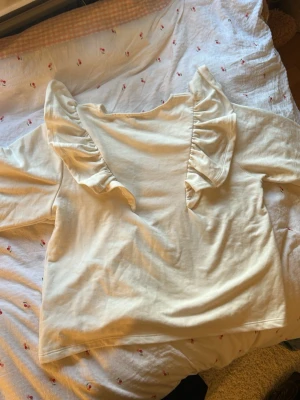 Vit topp med volangdetalj - Söt vit topp med långa ärmar och volangdetaljer längs axlarna. Toppen har en rund halsringning och är tillverkad i ett mjukt material som känns skönt mot huden. Perfekt för dig som gillar en romantisk och trendig stil.L men sitter som S/M