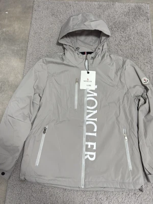 Grå vindjacka från Moncler - Snygg grå vindjacka från Moncler med stor logga längs dragkedjan framtill. Jackan har huva, tre dragkedjeförsedda fickor och Moncler-märke på ärmen. Tillverkad i lätt och slitstarkt material, perfekt för blåsiga dagar.
