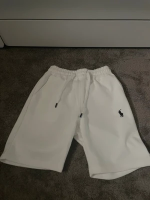 Vita shorts med snörning - Säljer ett par vita shorts med resår i midjan och snörning framtill. Shortsen har sidofickor och en liten broderad logga på vänster ben. Perfekta för sommaren och riktigt sköna att bära till vardags.