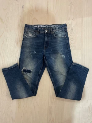 Blå slitna jeans från hoodrich - Säljer ett par blå jeans med slitningar och hål på ena benet. Jeansen har en rak passform, fem fickor och trycket 'FROM NOTHING TO SOMETHING' i midjan. Baksidan har två fickor och en liten broderad detalj. Perfekta för en avslappnad streetstil.