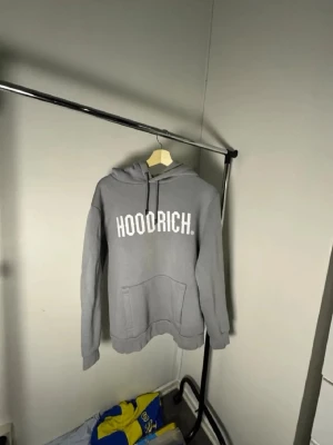 Grå HOODRICH hoodie med tryck - Snygg grå hoodie från HOODRICH med stor vit logga på bröstet. Klassisk modell med huva och dragsnören samt en stor magficka. Perfekt för dig som gillar streetwear och vill ha en enkel men stilren look.