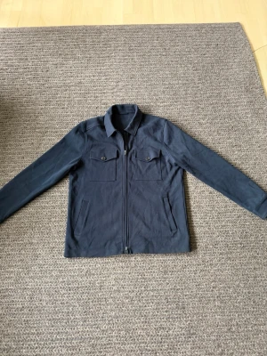 Mörkblå overshirt med dragkedja - Snygg mörkblå overshirt med dragkedja framtill och två bröstfickor med knappar. Overshirten har klassisk krage och raka ärmar. Materialet ser ut att vara kraftig bomull eller bomullsblandning, perfekt för lager på lager.