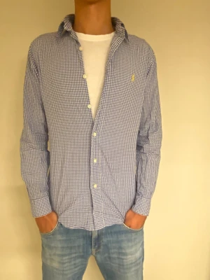 Blårutig skjorta från Polo Ralph Lauren - Säljer en blårutig skjorta från Polo Ralph Lauren i slim fit. Skjortan har klassisk krage, långa ärmar och knappar framtill. Tillverkad i mjuk bomull och har en liten gul broderad logga på bröstet. Perfekt för en clean och stilren look.