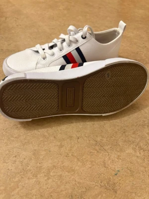 Vita Tommy Hilfiger sneakers - Snygga vita sneakers från Tommy Hilfiger med klassiska röda och blå ränder på sidan. Skorna har lågt skaft, vita snören och texten 'TOMMY' på hälen. Tillverkade i ett tåligt syntetmaterial med gummisula för bra grepp.