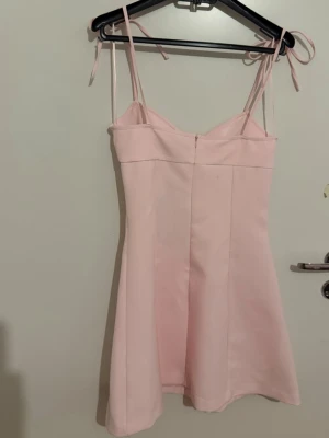 Ljusrosa kort klänning med knytband - Söt ljusrosa kort klänning med smala axelband som knyts upptill. Klänningen har en lätt utsvängd passform och en enkel, stilren design. Perfekt för varma dagar och somriga tillfällen.