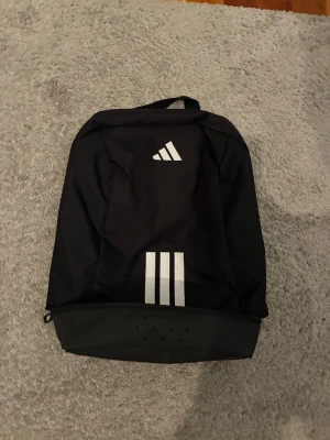 Svart ryggsäck från Adidas - Snygg svart skoväska från Adidas med klassiska vita ränder och logga framtill. Riktigt schysst helt ny aldrig använd