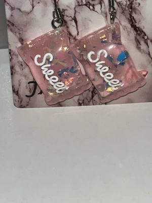 Rosa örhängen med texten 'Sweet' - Unika örhängen i form av rosa godispåsar med glitterdetaljer och vita texten  'Sweet' på framsidan. Tillverkade i resin med skimrande inslag som ger en lekfull och trendig look. Perfekta för dig som gillar färg och statement-accessoarer. Örhängens krokarna är i rostfritt stål 