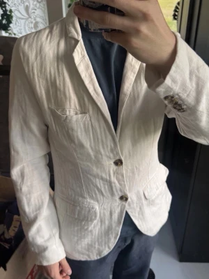 Vit linnékavaj från Lindbergh - Snygg vit kavaj i linne med två knappar framtill och klassisk krage. Kavajen har tre fickor fram och dekorativa knappar vid ärmsluten. Perfekt för en fräsch och stilren look. Passar till både jeans och chinos.