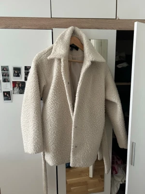 Beige teddyjacka med bälte - Säljer en beige teddyjacka med bred krage och knytskärp i midjan. Jackan har en fluffig, mjuk yta och stängs med tryckknappar framtill. Perfekt för dig som vill ha en mysig och trendig look under kyliga dagar. Modellen har vanligtvis S