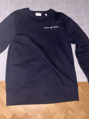 Svart sweatshirt från GANT - Klassisk svart sweatshirt från GANT med rund halsringning och tryckt logga samt '1949' på bröstet. Tröjan har ribbade muddar vid ärmslut och nederkant. Perfekt för en avslappnad och stilren look.