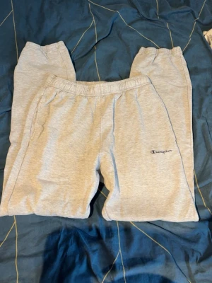Gråa mjukisbyxor från Champion - Säljer ett par ljusgrå mjukisbyxor från Champion med broderad logga på vänster ben. Byxorna har resår i midjan och två sidofickor. Perfekta för chill dagar eller när du vill vara extra bekväm. Klassisk passform och mjukt material.
