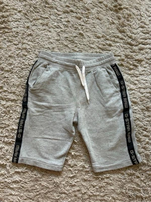 Grå shorts med snörning från G-Star RAW - Snygga grå shorts från G-Star RAW med svarta sidor där loggan står tryckt. De har snörning i midjan och två fickor fram. Perfekta för en avslappnad stil och sköna att bära under varma dagar.
