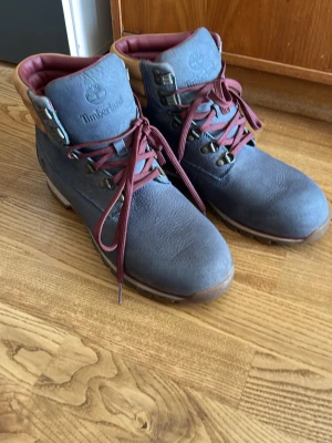 Blåa Timberland stövletter - Säljer ett par blåa stövletter från Timberland med vinröda snören och detaljer i beige runt ankeln. Skorna har robust sula och metallhål för snörningen. Tillverkade i slitstarkt läder och har en klassisk outdoor-look.