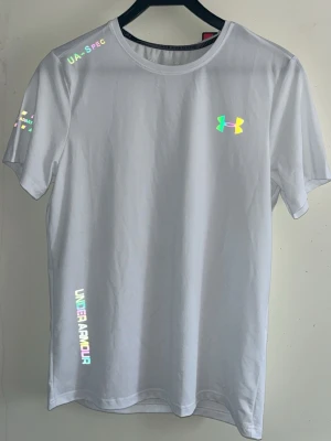 Vit Under Armour t-shirt med holografiska detaljer - Snygg vit t-shirt från Under Armour med holografiska loggor och textdetaljer på bröstet, axeln och sidan. Tillverkad i ett lätt och mjukt material som passar perfekt till träning eller chill. Klassisk rund halsringning och korta ärmar för en clean look.