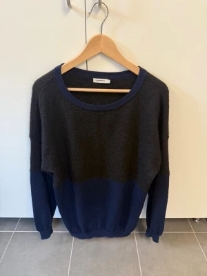 Mörkblå och svart stickad tröja J.Lindeberg - Snygg stickad tröja från J.Lindeberg i storlek M. Tröjan har en cool colorblock-design med mörkblå nederdel och muddar samt svart överdel. Rund halsringning och långa ärmar. Perfekt för dig som gillar stilrena plagg med en twist.