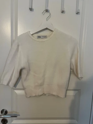 Vit stickad tröja från Zara - Säljer en enkel och stilren vit stickad tröja från Zara. Tröjan har rund halsringning, halvlånga ärmar och ribbade muddar vid ärmslut och nederkant. Perfekt att matcha med jeans eller kjol för en clean look.