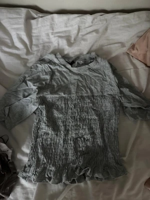 Grå ribbad topp med volang - Grå topp med ribbad struktur och volangkant nertill. Toppen har rund halsringning och långa ärmar. Perfekt för dig som gillar en enkel men ändå söt stil.