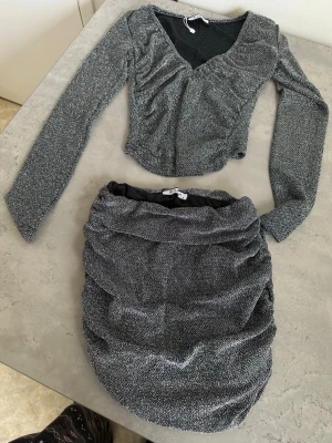 Glittrig croppad topp från X-tayn - Säljer en glittrig croppad topp från X-tayn i svart och silver. Toppen har lång ärm, v-ringning och rynkade detaljer framtill. Perfekt för dig som vill sticka ut lite extra med en snygg och trendig look.