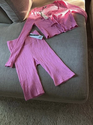 Rosa ribbat set - Säljer ett rosa ribbat loungewear-set med långärmad topp och matchande byxor. Toppen har knytning framtill och byxorna har en bekväm, hög midja. Materialet är mjukt och stretchigt, perfekt för chill dagar hemma.