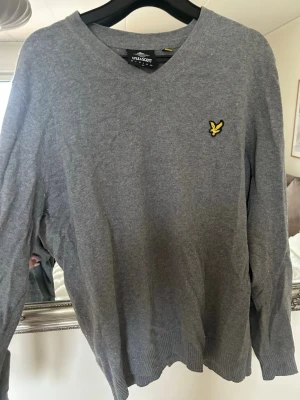 Grå stickad tröja från Lyle & Scott - Snygg grå stickad tröja från Lyle & Scott med V-ringning och den klassiska gula logon på bröstet. Tröjan har långa ärmar och ribbade muddar vid ärmslut och nederkant. Perfekt till jeans eller chinos för en clean look.