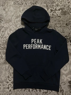 Mörkblå hoodie Peak Performance - Snygg mörkblå hoodie från Peak Performance med stor vit logga på bröstet. Klassisk modell med huva och dragsko samt en stor magficka. Perfekt för en avslappnad och sportig stil. Mjuk och skön bomullsblandning.