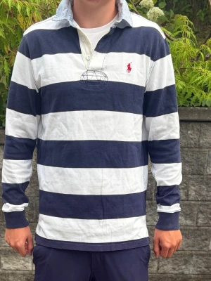 Randig rugbytröja från Polo Ralph Lauren - Säljer en långärmad rugbytröja från Polo Ralph Lauren med breda marinblå och vita ränder. Tröjan har krage i ljus färg, knäppning vid halsen och den  röda loggan broderad på bröstet. 
